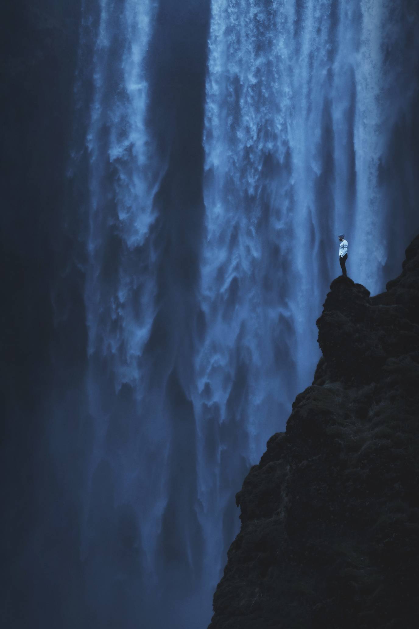 A person in a white sweater stands next to Skogafoss Waterfall in Iceland