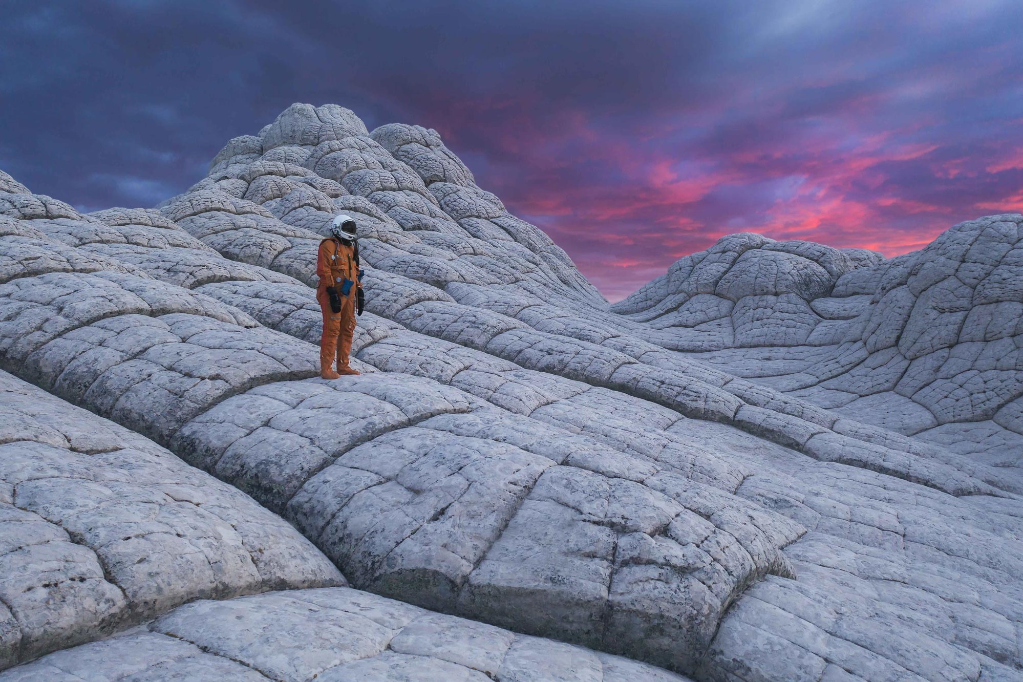 An astronaut wearing an orange space suit stands in the desert in Arizona with a beautiful, pink sky sunset at White Pocket.