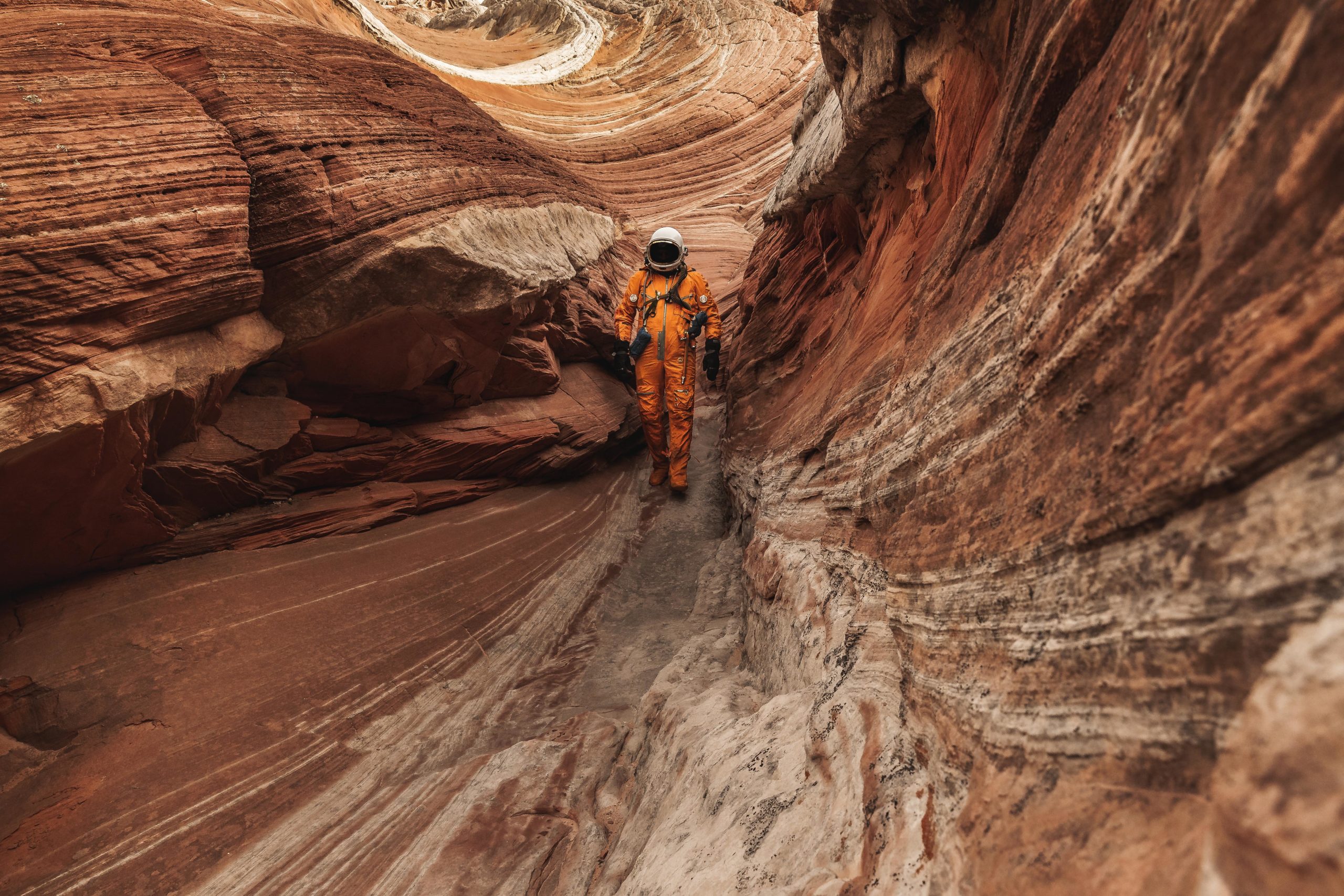 Photography of an astronaut wearing on orange space suit walking through a slot canyon in the otherworldly and mars-like landscape of Arizona, USA.