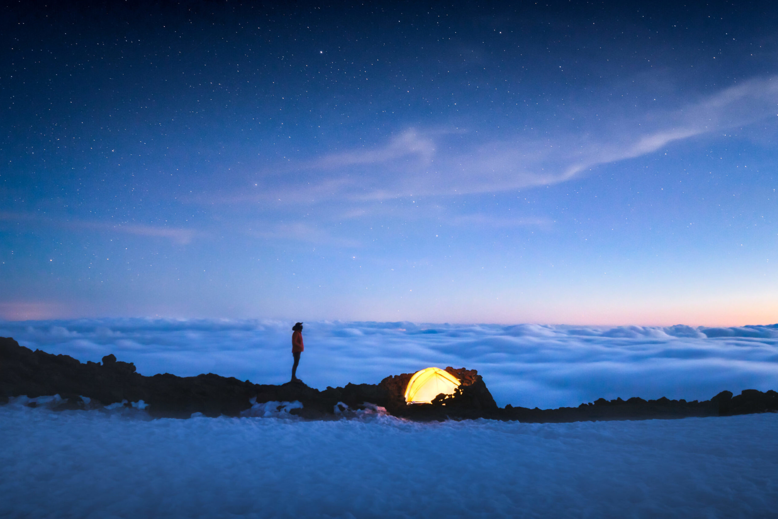 A tent glows yellow under a starry night on the Summit of South Sister mountain in Oregon above the fog. Photo by Taylor Gray Visuals